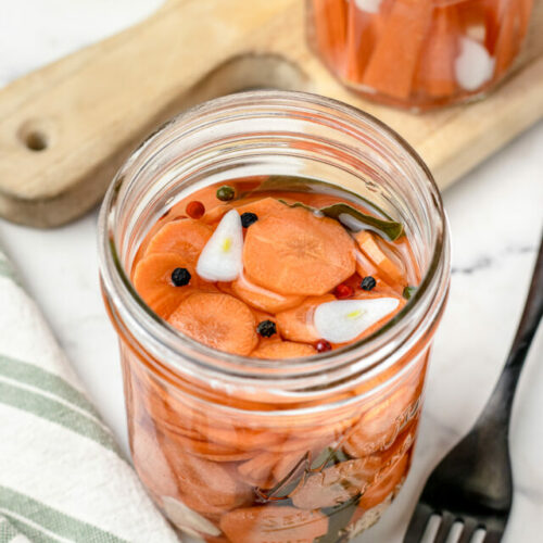 Jar of pickled carrots with fork on the side for eating. Another jar of pickled carrot sticks in the background.