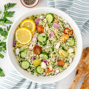 Lemon Orzo Salad with Feta, fresh vegetables and herbs topped with a lemon dressing, black pepper, and a few lemon slices. Spoons for tossing and serving the salad on the side.