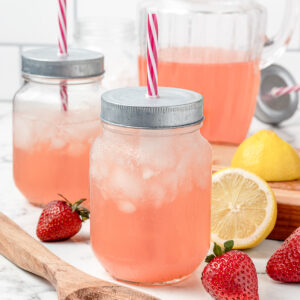 2 jars of Pink Lemonade with fresh strawberries and lemon on the side. There is a large pitcher of more lemonade in the background.