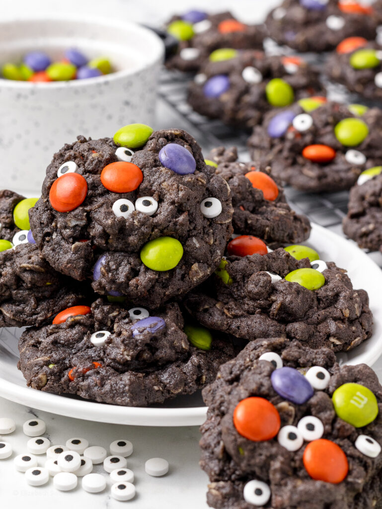 Plate of chocolate Halloween Monster Cookies with candy eyes and Halloween colored M&M's. A cooling rack of more cookies on the side.