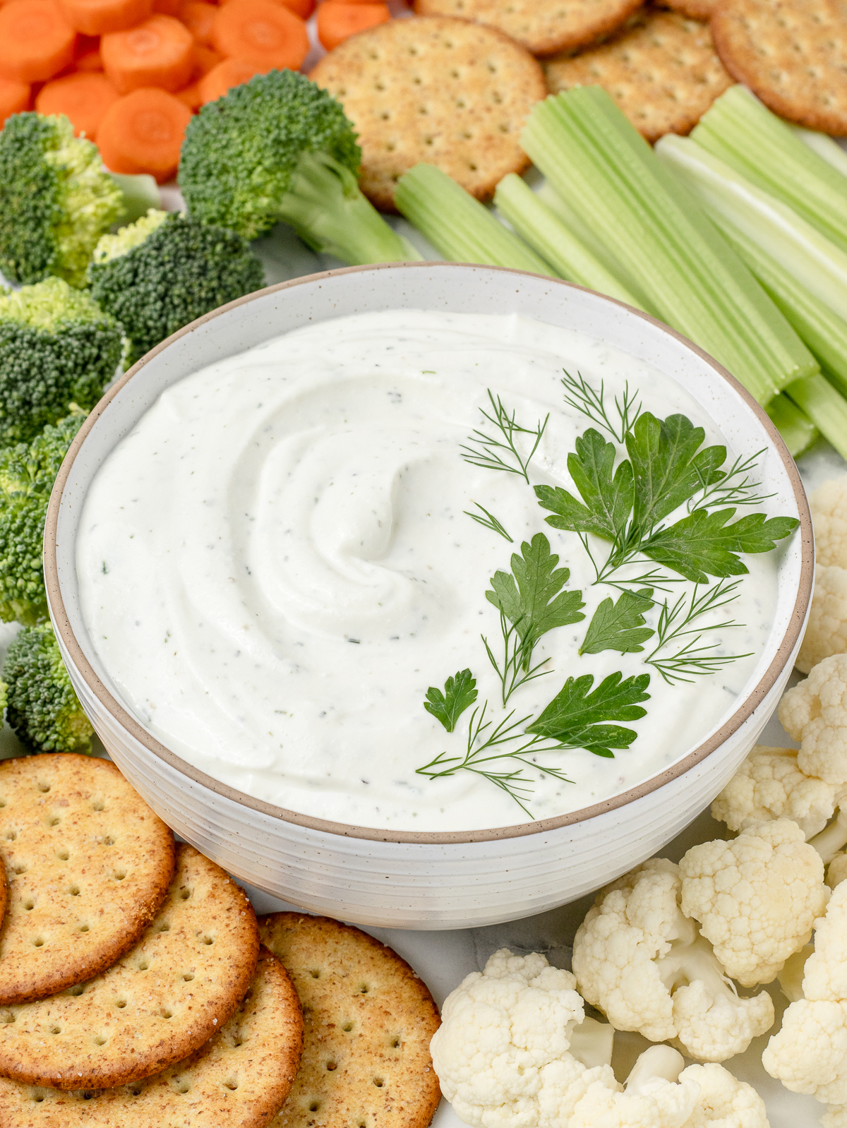 Bowl of ranch dip topped with fresh parsley and fresh dill. There are crackers, cauliflower, broccoli, celery, and carrots on the side.