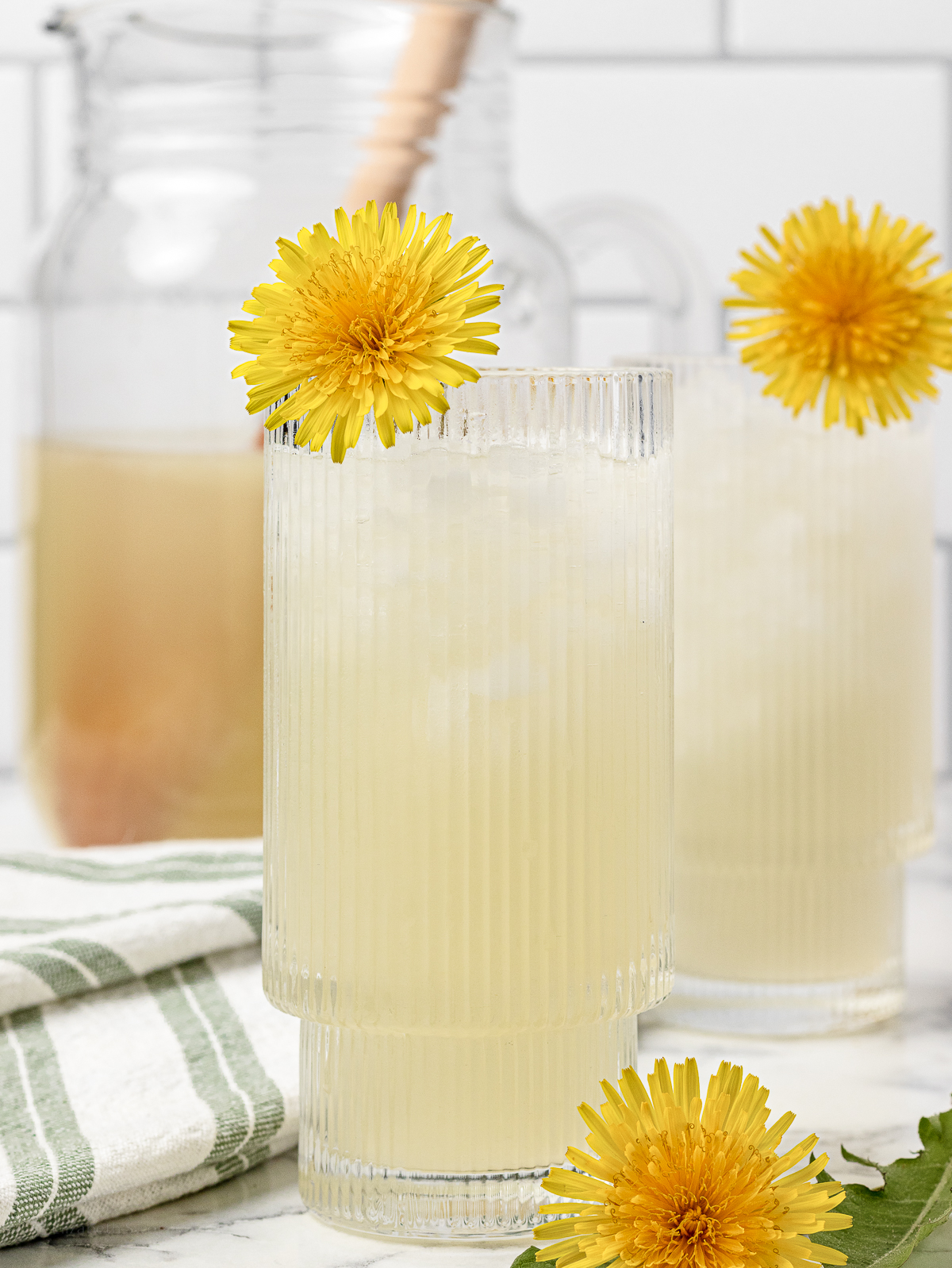 Two glasses of iced Dandelion Lemonde garnished with a dandelion flower. There is a pitcher of more in the back for pouring more.