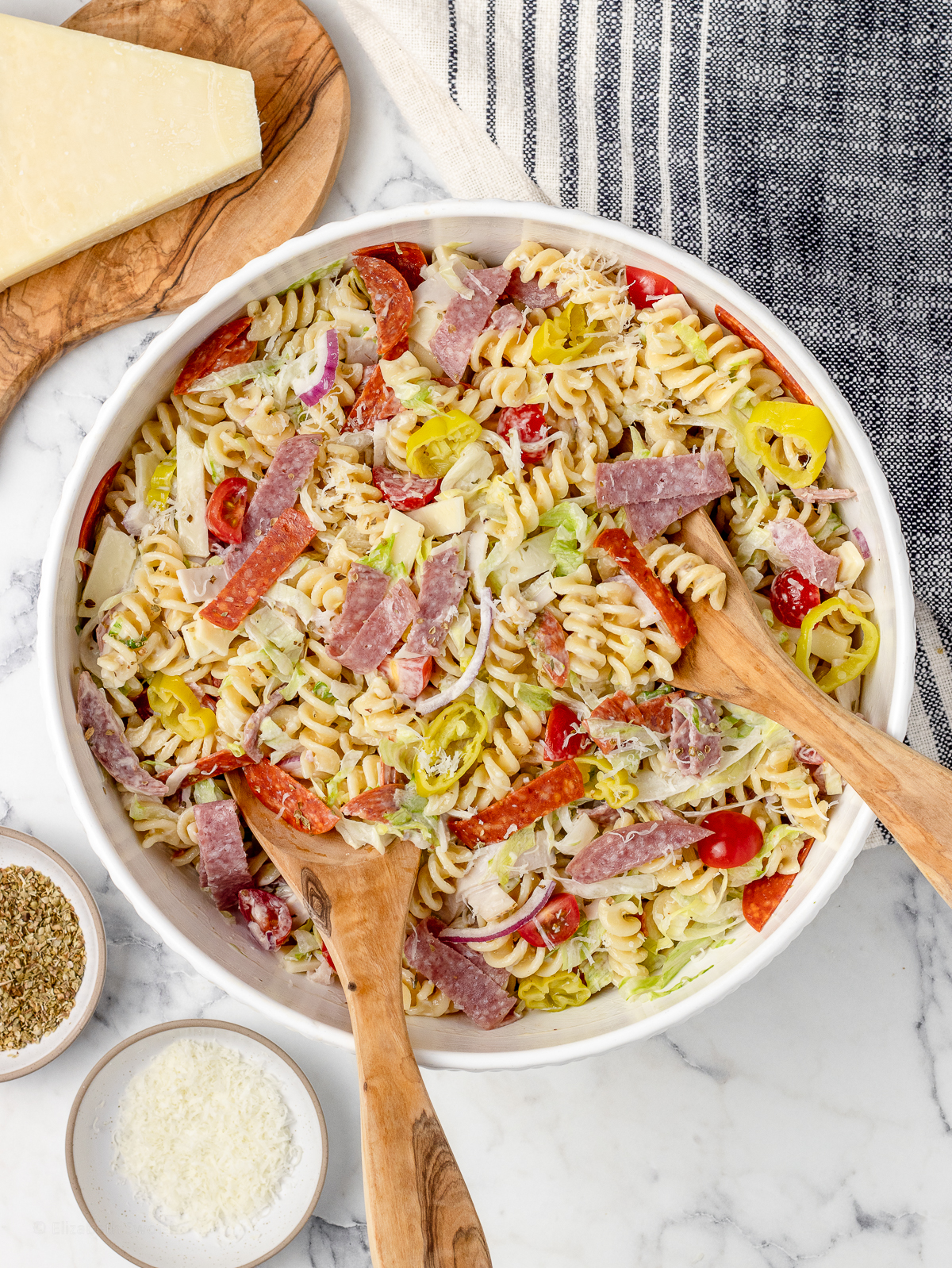 Overhead image of the pasta salad with 2 serving spoons in it. There is more dried oregano and freshly grated parmesan cheese on the side.