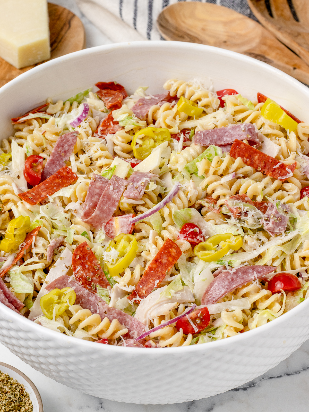 A big bowl of Grinder Pasta Salad with serving spoons, dried oregano, and extra parmesan cheese on the side