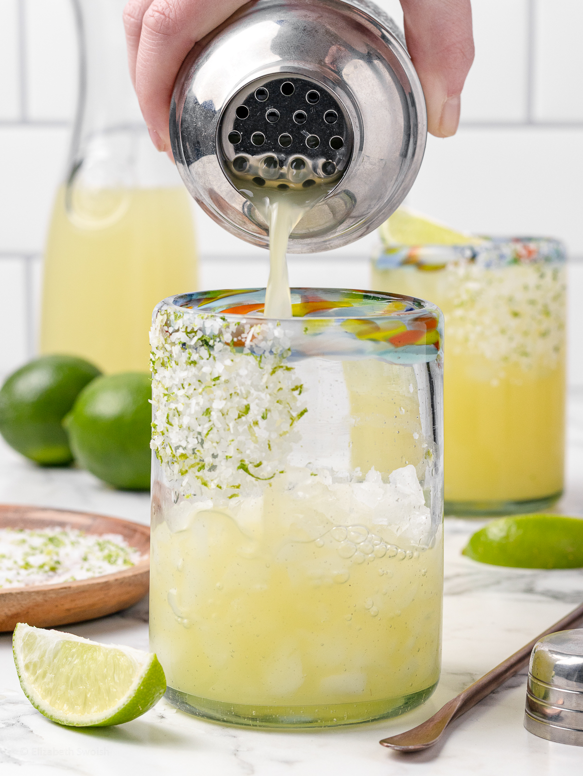 Pouring the drink into an ice filled glass. The glass is garnished with flaky sea salt and lime zest. There are lime slices, and a plate of the salt zest mixture.