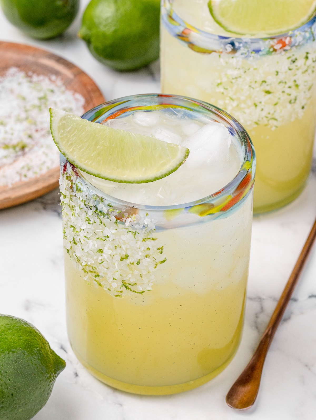 A non alcoholic margarita with a lime wedge on top. There is another drink in the background and limes all around.