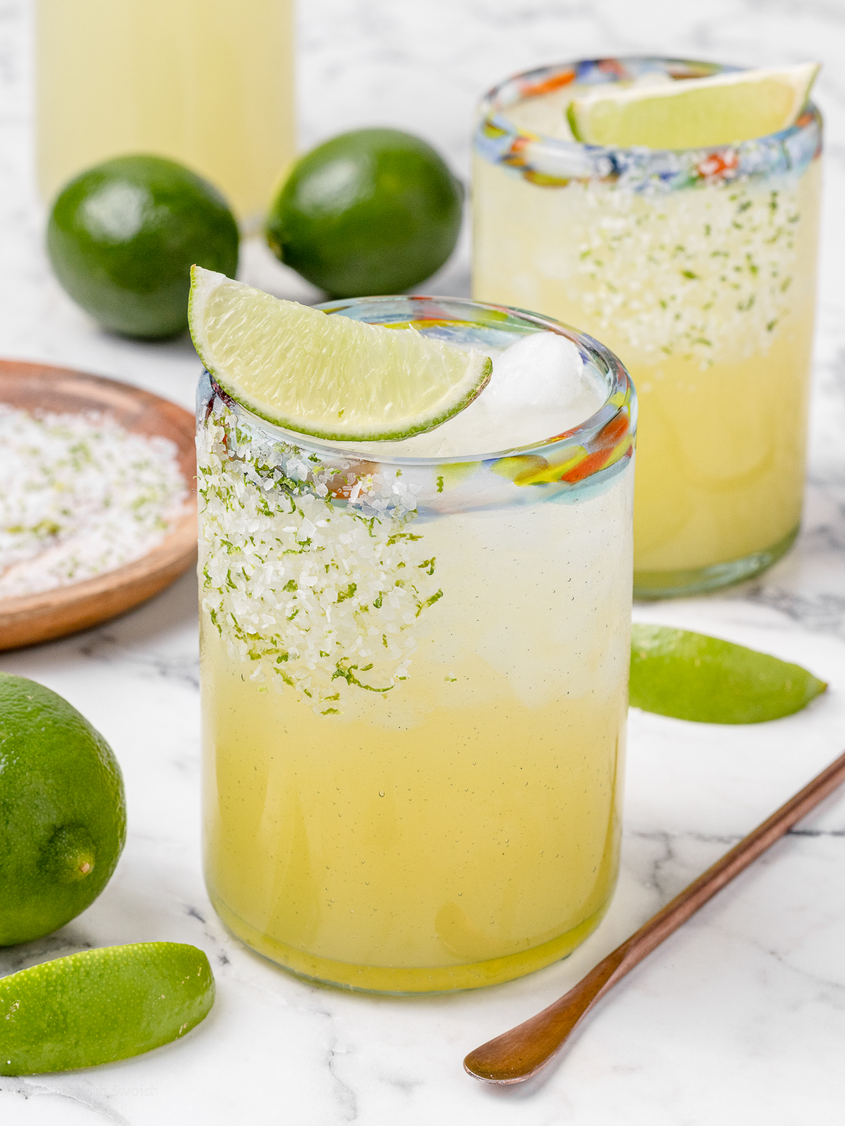 Two non alcoholic margarita mocktails and a pitcher with more in the background. There is also limes, lime wedges, and a plate of salt and lime zest for garnishing the rim.