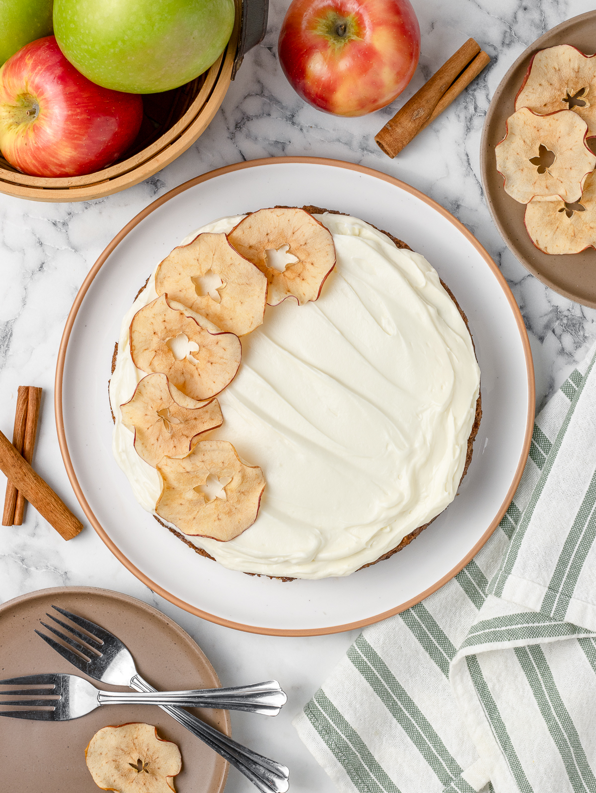 Overhead image of the cake. There is plates, forks, dried apples, red apples, green apples, and cinnamon sticks around it.