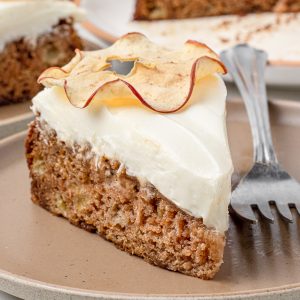Slice of Apple Spice Cake sitting on a plate. It is has a layer of cream cheese frosting and a dried apple slice on top. There are bits of apple in the cake as well as flecks of fall spices.