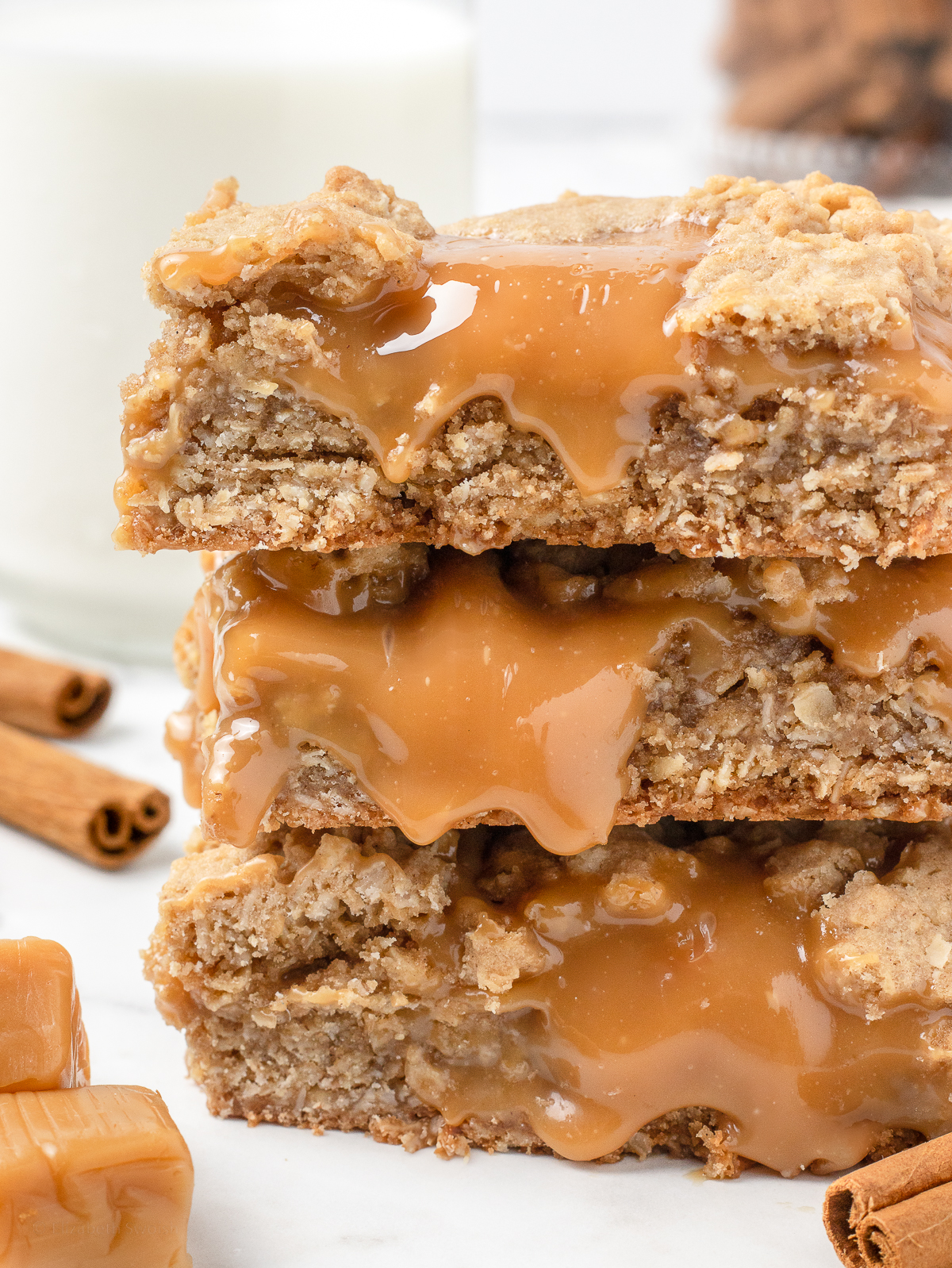 Close up of the bars to see their gooey caramel layers. There is a glass of milk, cinnamon sticks, and caramel squares in the background.