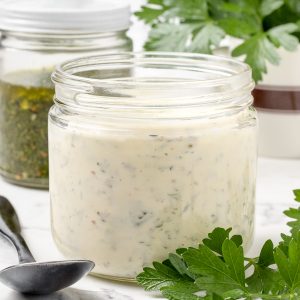 Jar of Chimichurri Aioli sauce ready for serving. There is a spoon, a jar of chimichurri, and more fresh parsley surrounding it.