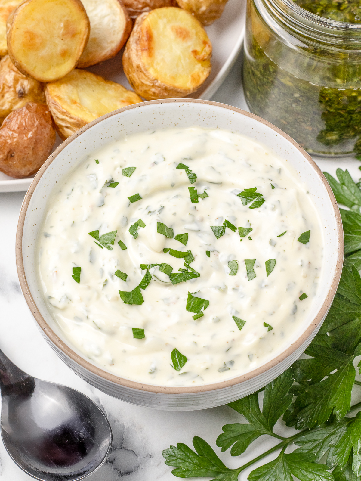 Chimichurri Aioli sauce in a bowl with a spoon on the side for serving. There is also a jar of chimichurri, fresh parsley, and roasted potatoes for dipping.