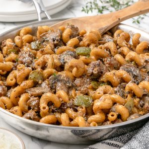 A pan of Creamy Italian Sausage Pasta with mushrooms and green peppers. There is a spoon for serving, 2 plates and forks plus fresh thyme and freshly grated parmesan cheese.