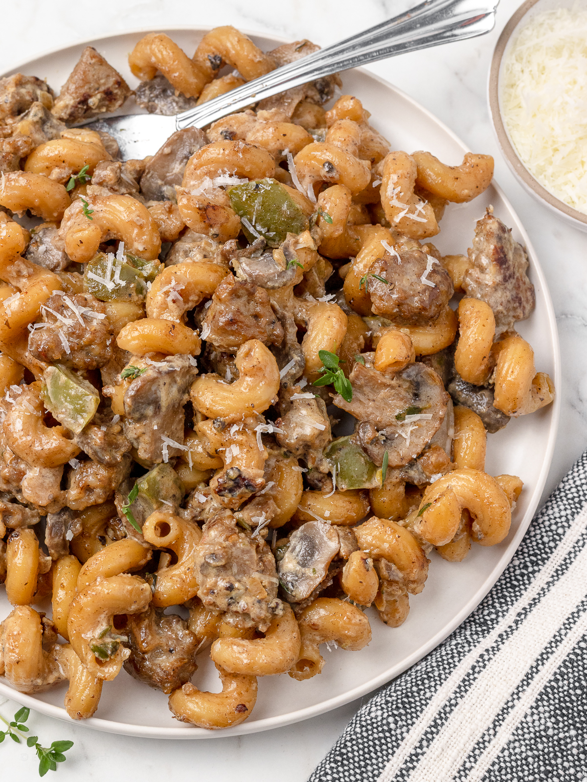 Plate of pasta with parmesan cheese on the side. There is a fork for eating the creamy pasta with Italian sausage, green peppers, and mushrooms.