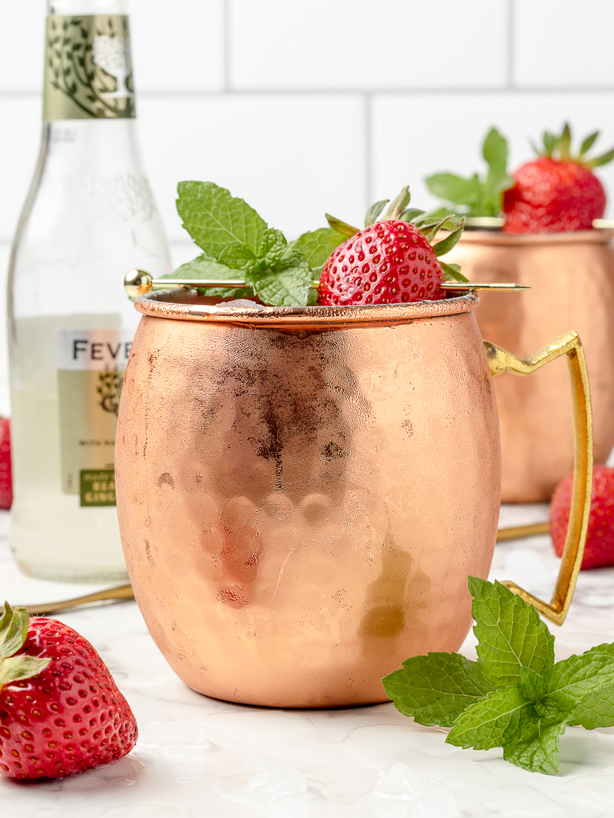 A fresh Strawberry Mule Mocktail surrounded by strawberries, mint leaves, more ginger beer, and spoon for stirring.