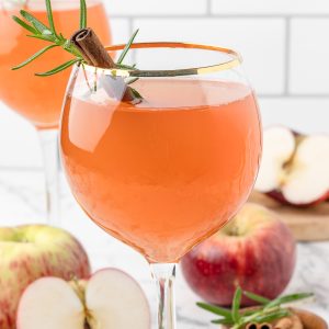 Apple Cider Aperol Spritz Mocktail in a gold rimmed wine glass with a cinnamon stick and rosemary garnish. There is another glass of it in the background with apples scattered around.
