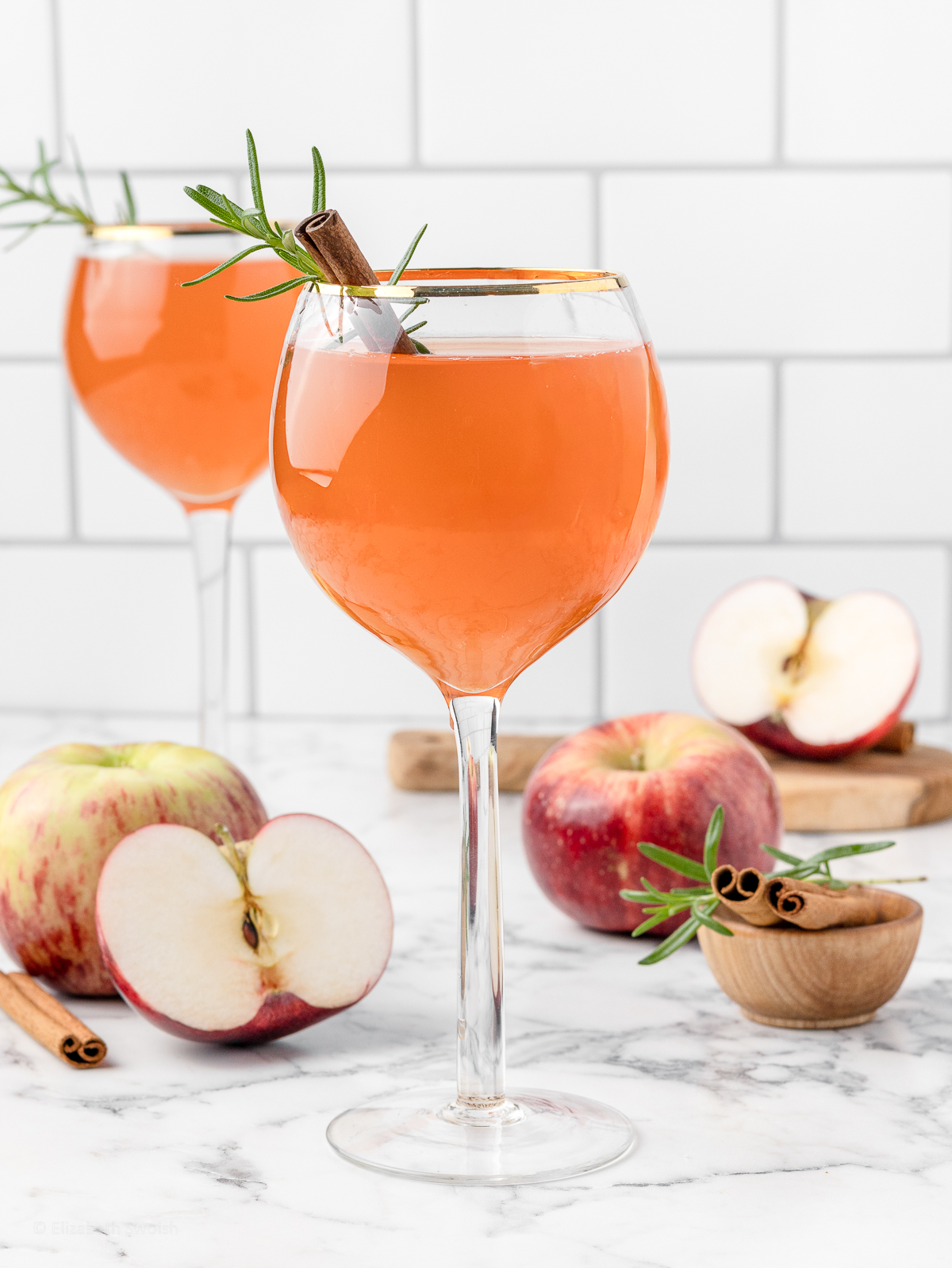 Two light orange fall aperol spritz mocktails with apples, rosemary, and cinnamon sticks all around.