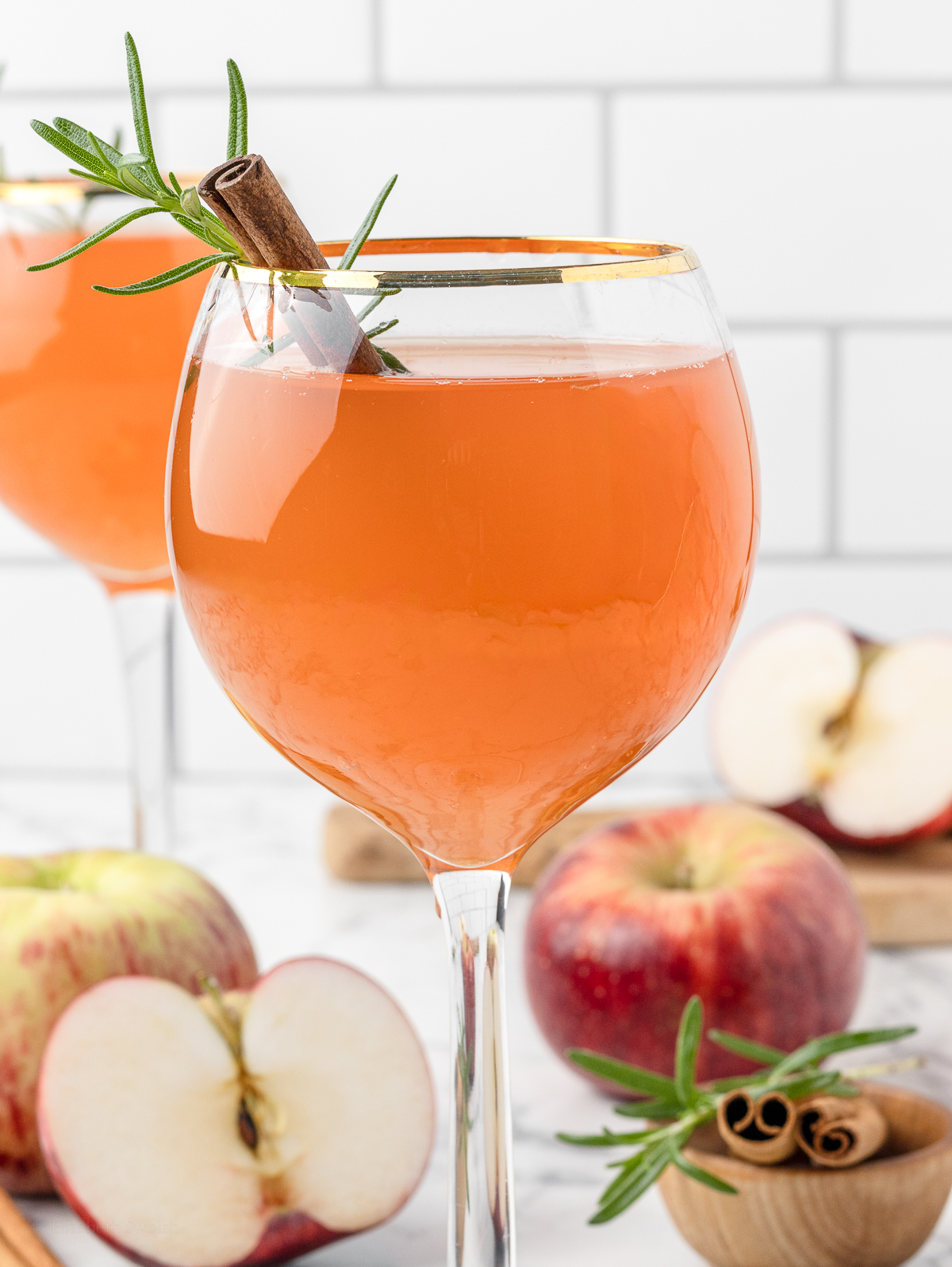 Apple Cider Aperol Spritz Mocktail in a gold rimmed wine glass. There is another in the background with apples, rosemary, and cinnamon sticks scattered around.