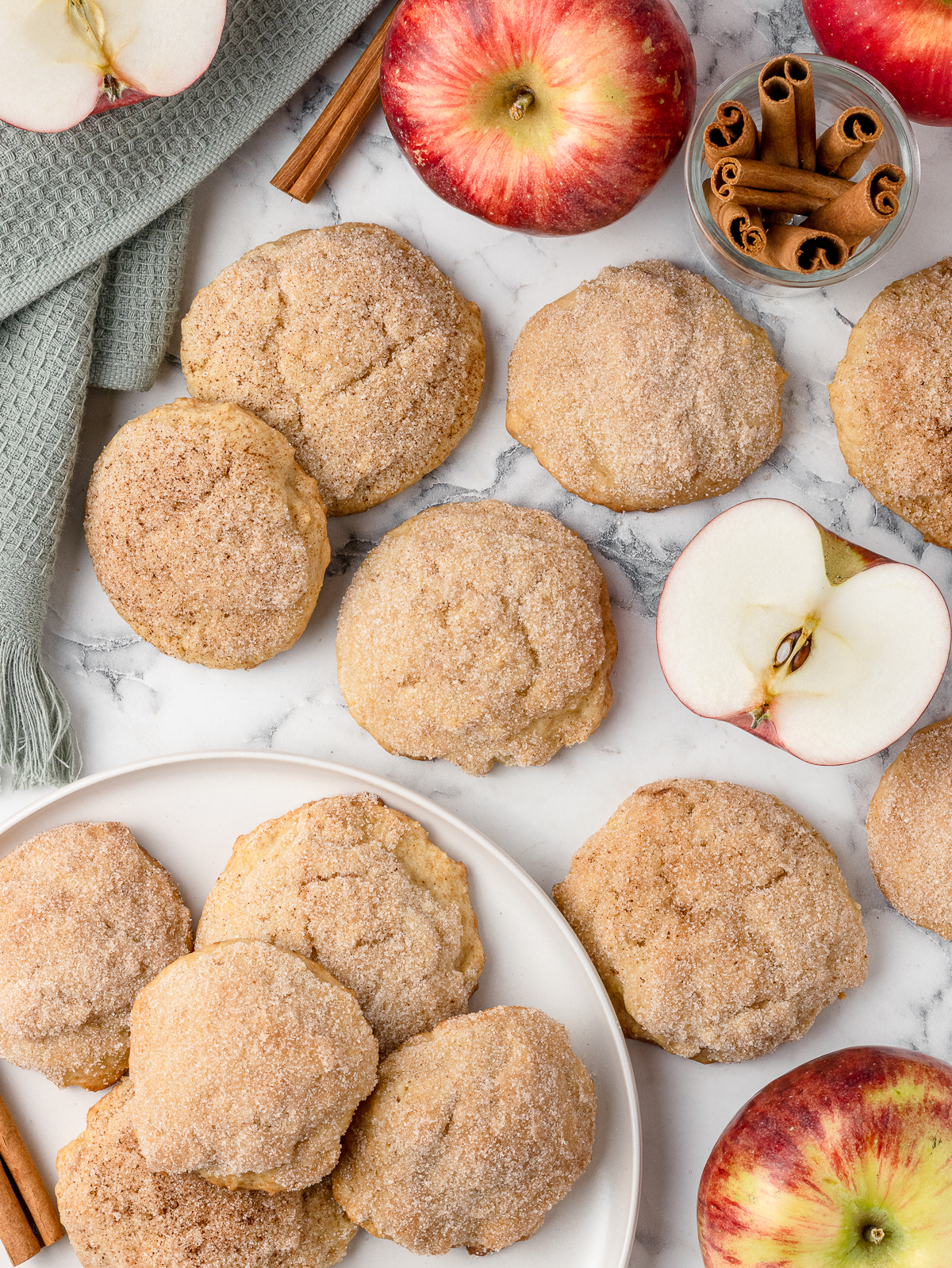 Overhead view of cookies on a plate and scattered around it. You can see the cinnamon sugar tops and there are apples and cinnamon sticks on the side.