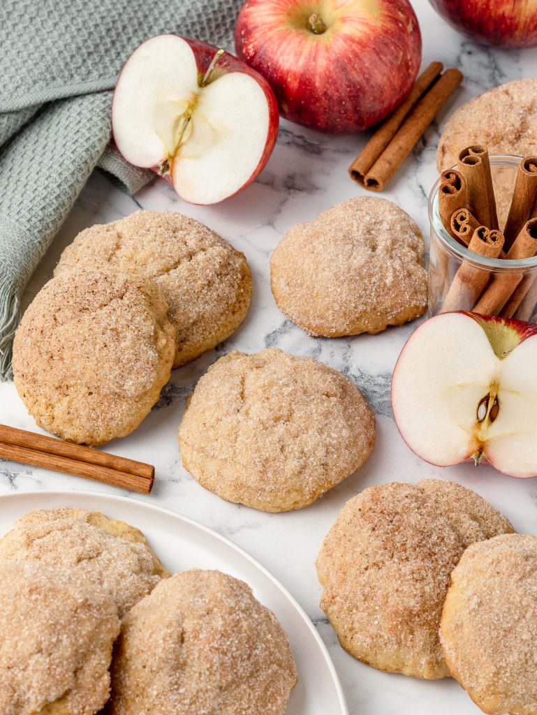 Cookies on a plate and scattered around it. You can see the cinnamon sugar tops and how thick and soft the cookies are. There are apples and cinnamon sticks around them.