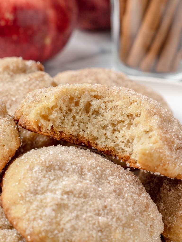 Big bite taken from the cookie. It shows a soft, thick texture with cinnamon sugar topping. There are more cookies under it with apples and cinnamon sticks behind it.