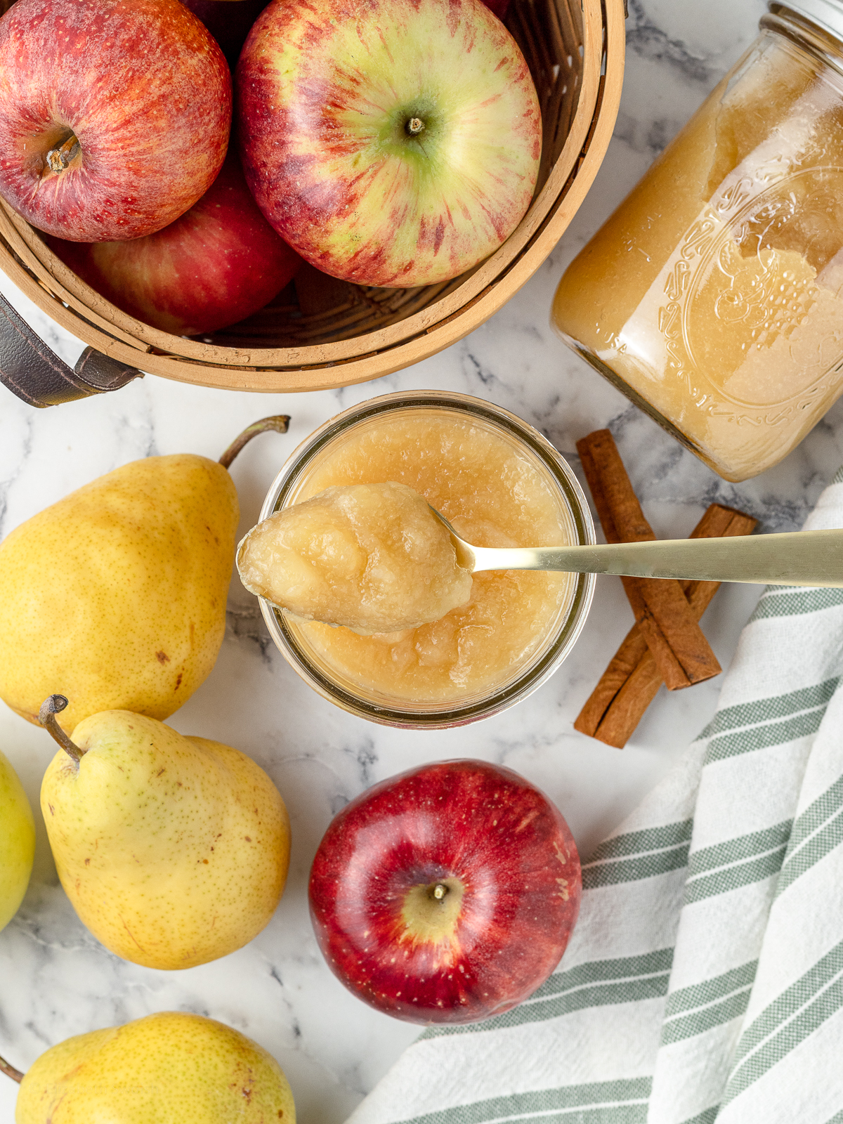 A spoon of pear applesauce sitting on top of the jar. More fresh apples and pears plus cinnamon sticks surround it.