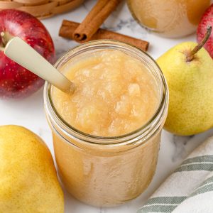 Smooth, cinnamon spiced Pear Applesauce in a jar. There is a spoon in it for serving and more fresh apples, pears, and cinnamon sticks surrounding it.