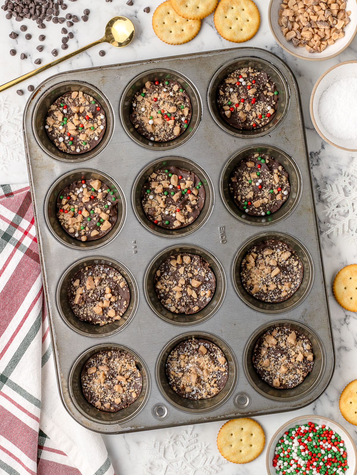 Cookies in the baking pan surrounded by more of the ingredients; Ritz crackers, chocolate chips, sprinkles, toffee pieces, and flaky sea salt.