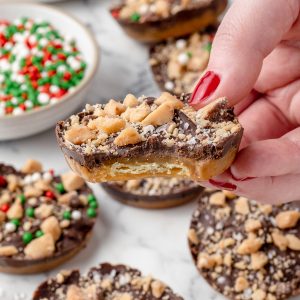 Hand holding a cookie with a bite taken from it, you can see the layers of cracker, toffee, and chocolate. There are more Christmas Crack Cookies in the background.