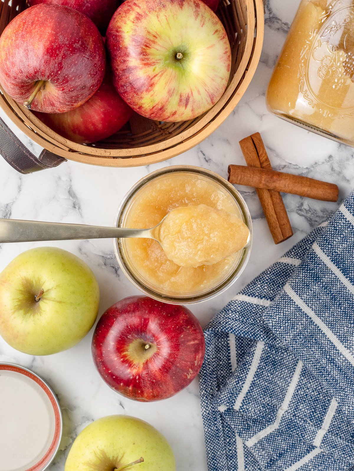 Cinnamon applesauce surrounded by a variety of apples, cinnamon sticks, and another jar of it. There is a spoon resting on it filled with the applesauce.