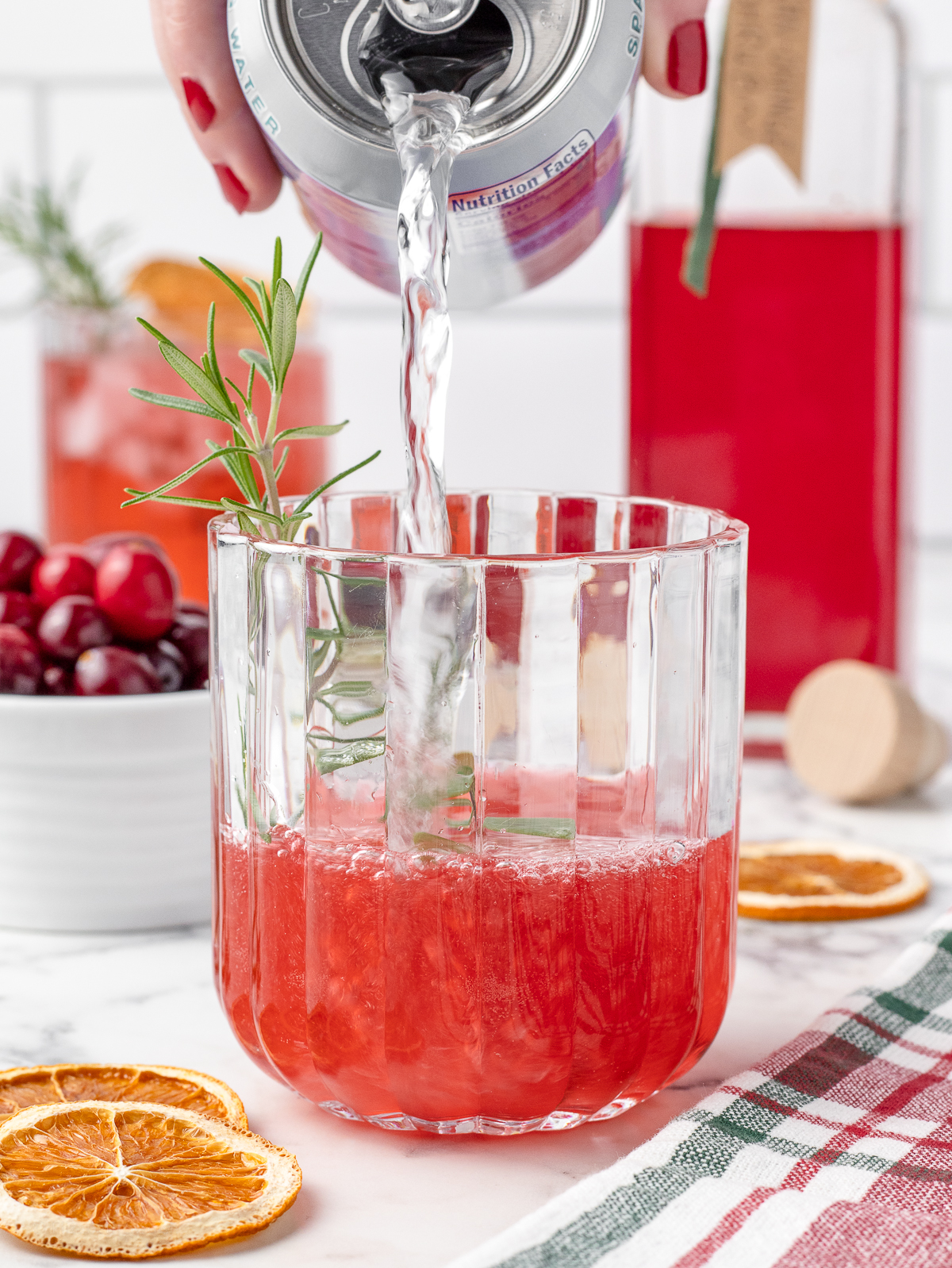 Sparkling water being poured over the shrub. There is rosemary, fresh cranberries, and dried orange slices for garnishing and pears in the background.