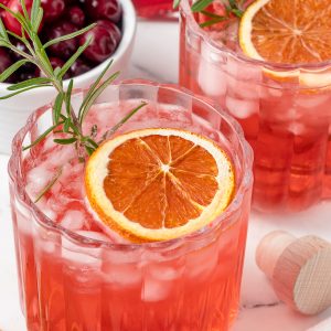 Close up of Cranberry Shrub drink garnished with orange slice and rosemary. There is another shrub and bowl of fresh cranberries in the back.