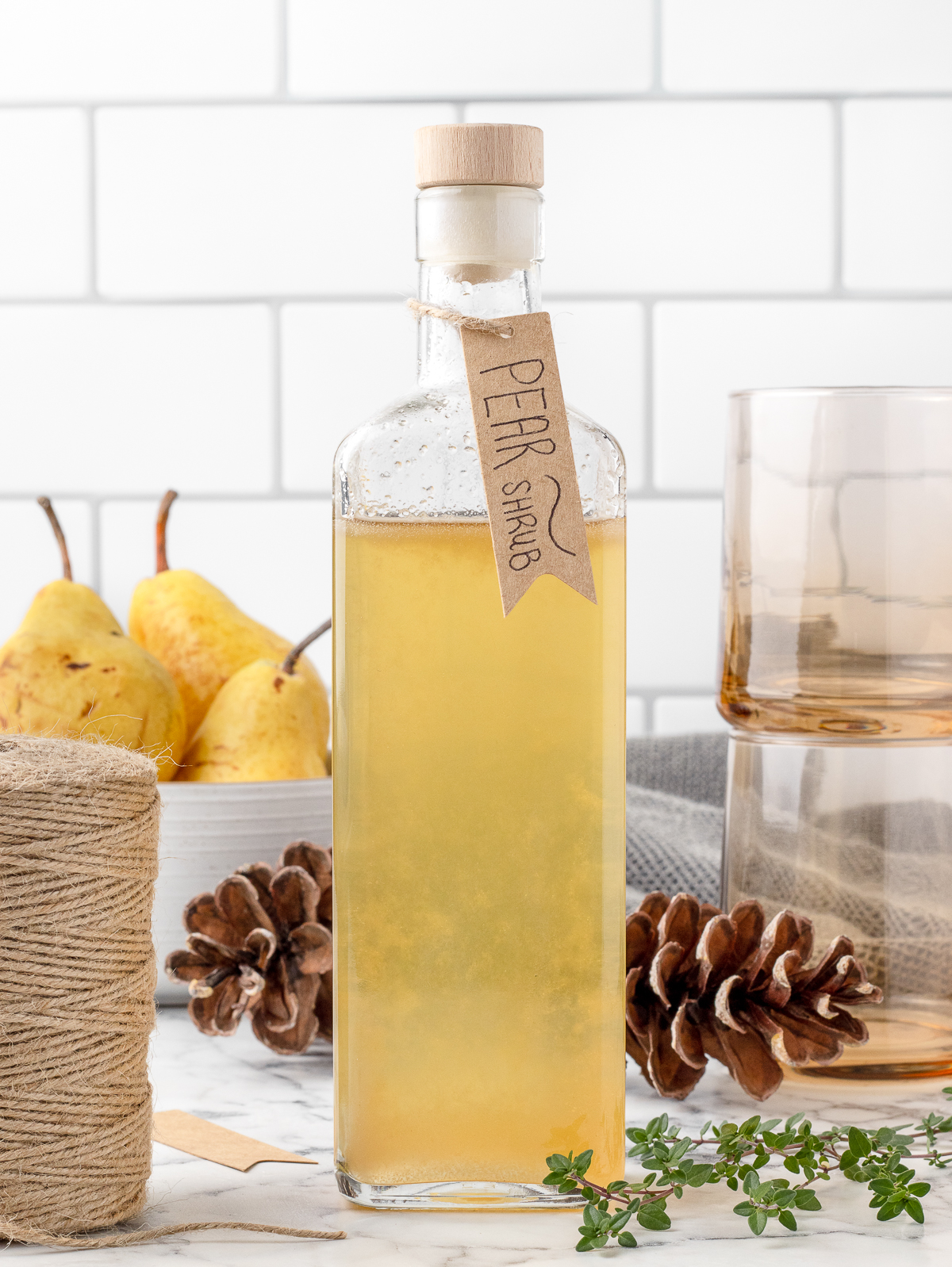 Pear Shrub in a bottle with a gift tag. There's twine, glasses, pears, and thyme on the side.