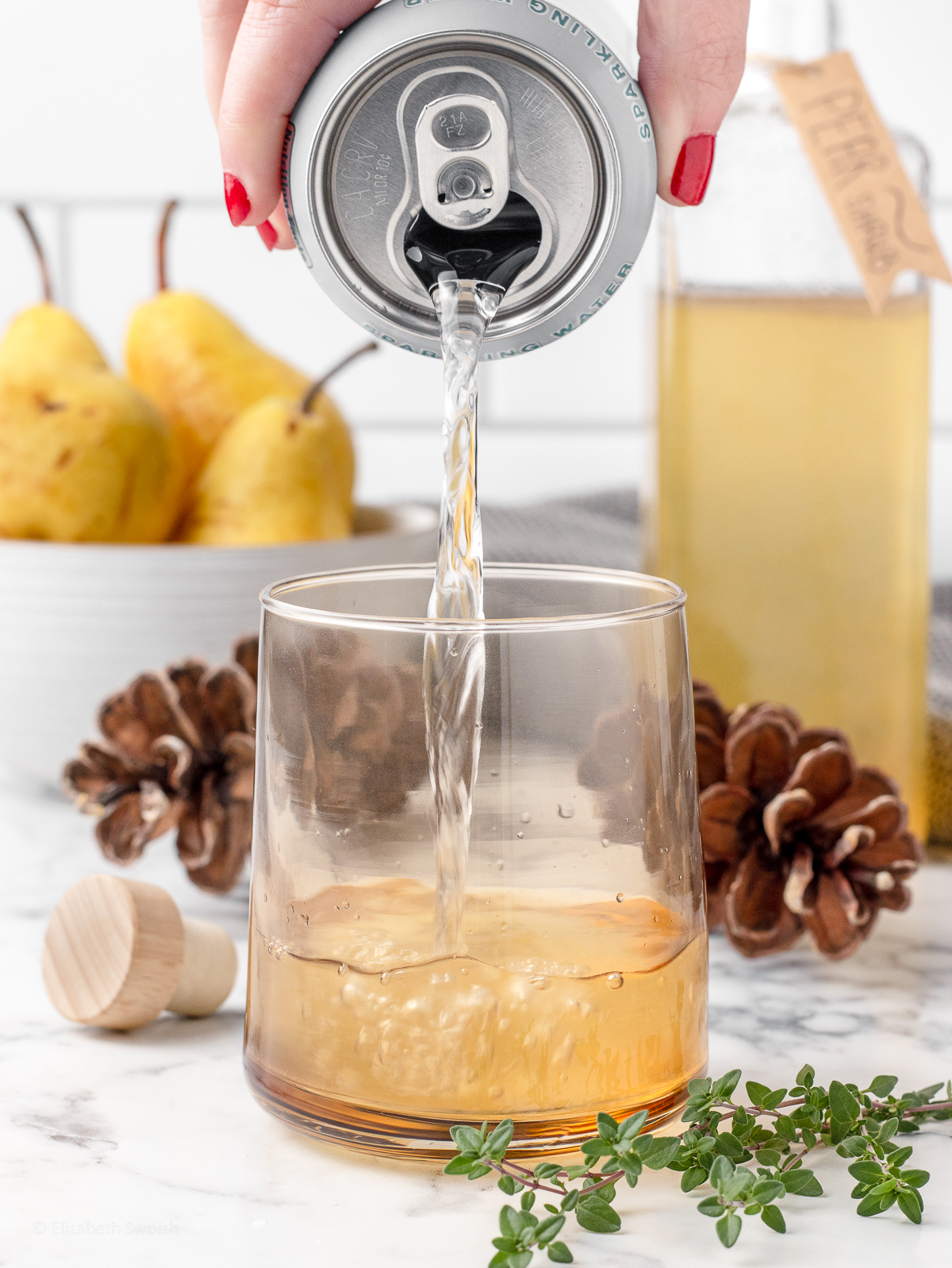 Sparkling water being poured over the shrub. There is thyme for garnishing and pears in the background.