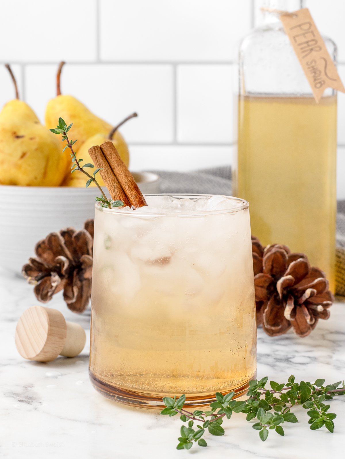 Pear Shrub drink in a glass garnished with a sprig of thyme and a cinnamon stick. There is more shrub in a bottle in the background and fresh pears.