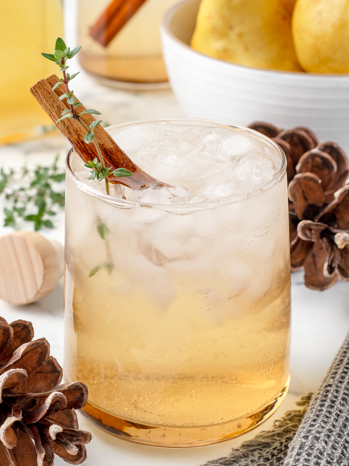 A shrub drink garnished with thyme and cinnamon stick with a bowl of pears in the background.