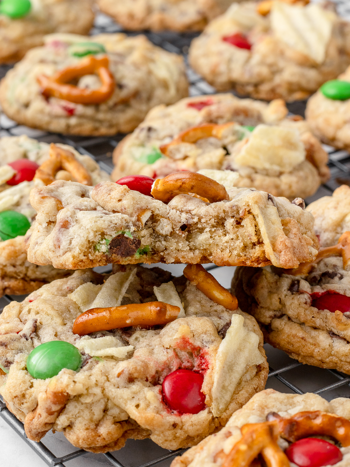 Christmas Kitchen Sink Cookies on a cooling rack with one that has a bite taken from it. You can see red and green colored candies, wavy potato chips, pretzels, toffee bits, and chocolate chips.