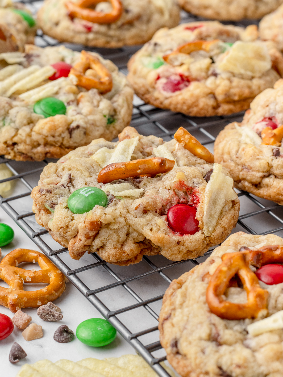 Cookies on a cooling rack. You can see how they have red and green colored candies, wavy potato chips, pretzels, toffee bits, and chocolate chips inside them.