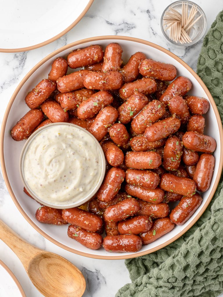 Overhead image with sausages on a plate and a dipping sauce on the side. There is a spoon for serving or toothpicks for grabbing them one by one.