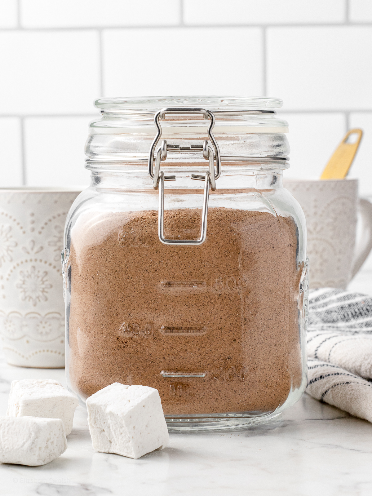 Big jar filled with Hot Chocolate Mix and mugs and marshmallows on the side.
