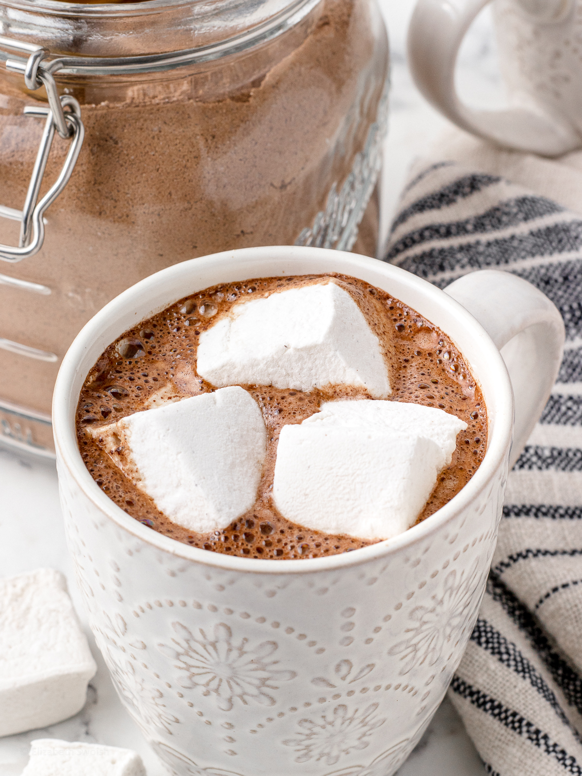 Cup filled with Hot Chocolate and topped with homemade marshmallows. There is a jar of the mix in the background.