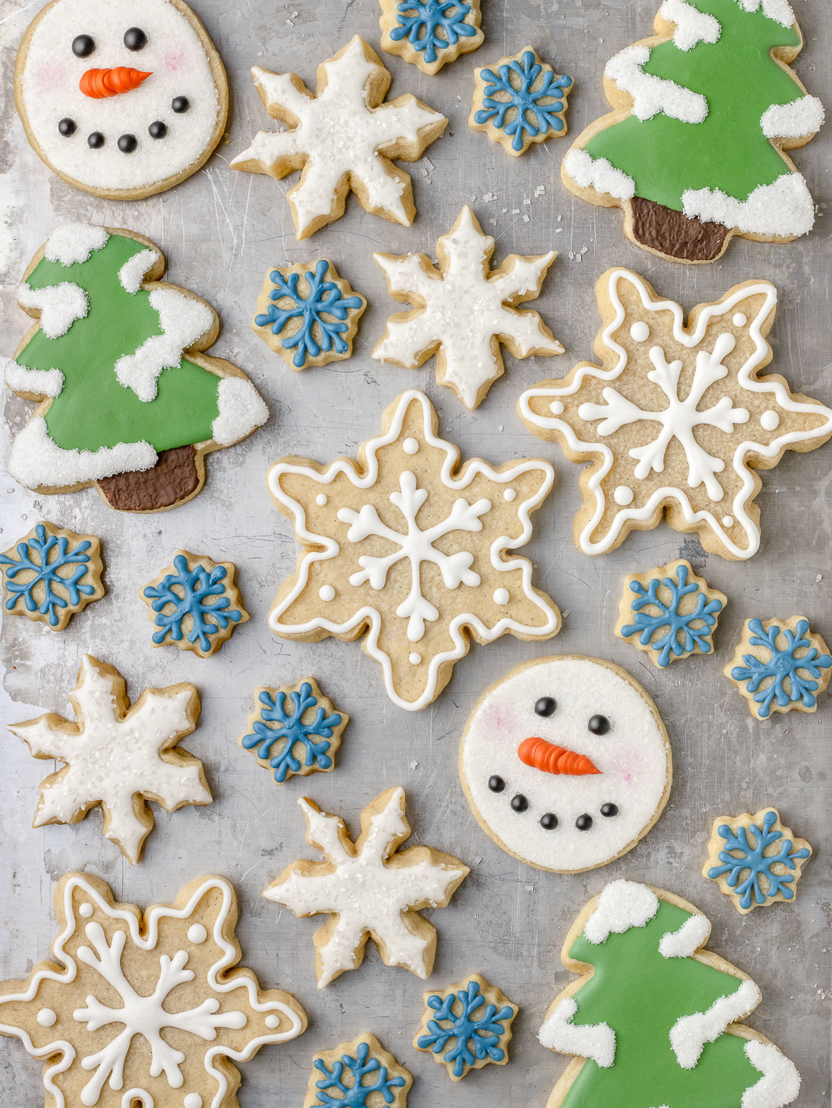 Scattered Vanilla Sugar Cookies. There are some decorated snowflakes iced with sprinkles, green pine trees with snow, and cute snowman faces with rosy cheeks.