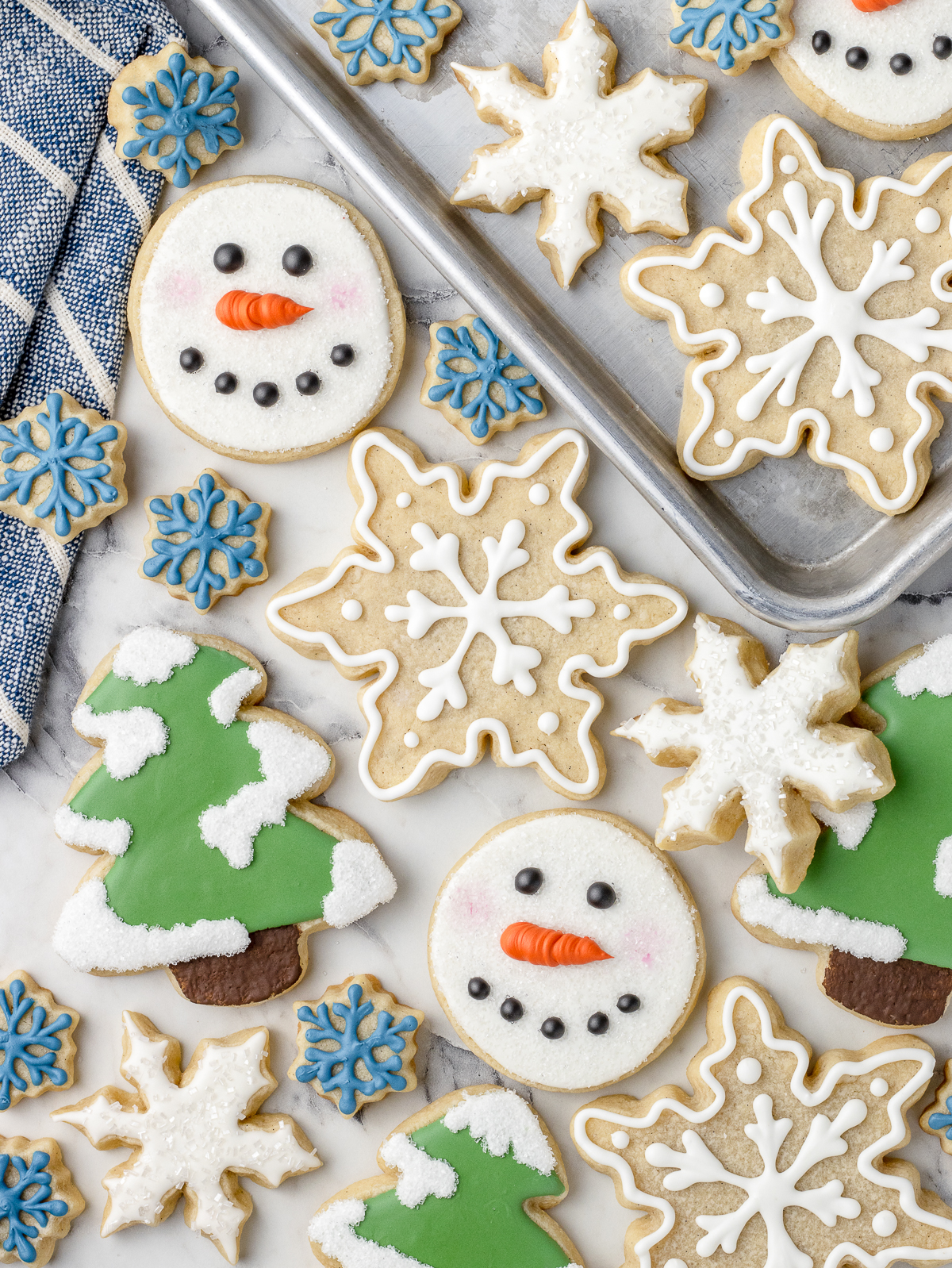 Scattered sugar cookies. There are decorated snowflakes iced with sprinkles, pine trees with snow, and cute snowman faces.