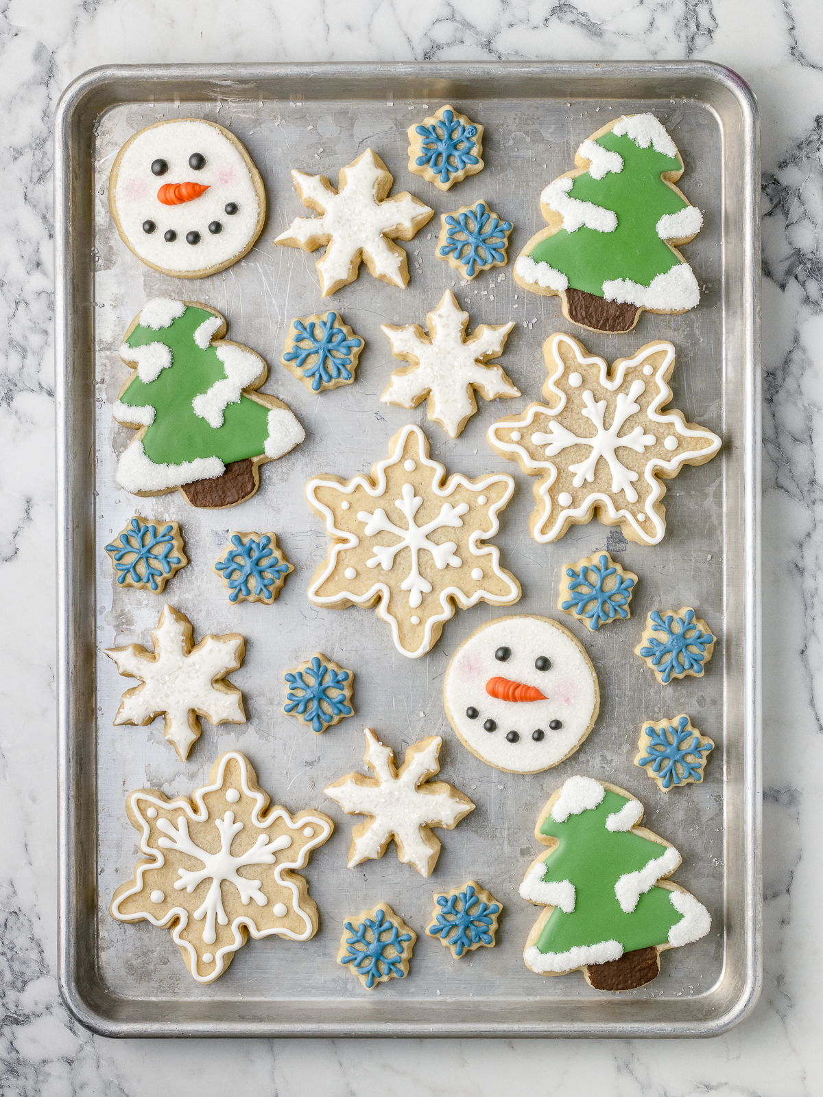 Decorated sugar cookies on a sheet pan. There are snowflakes iced with sprinkles, pine trees with snow, and cute snowman faces.