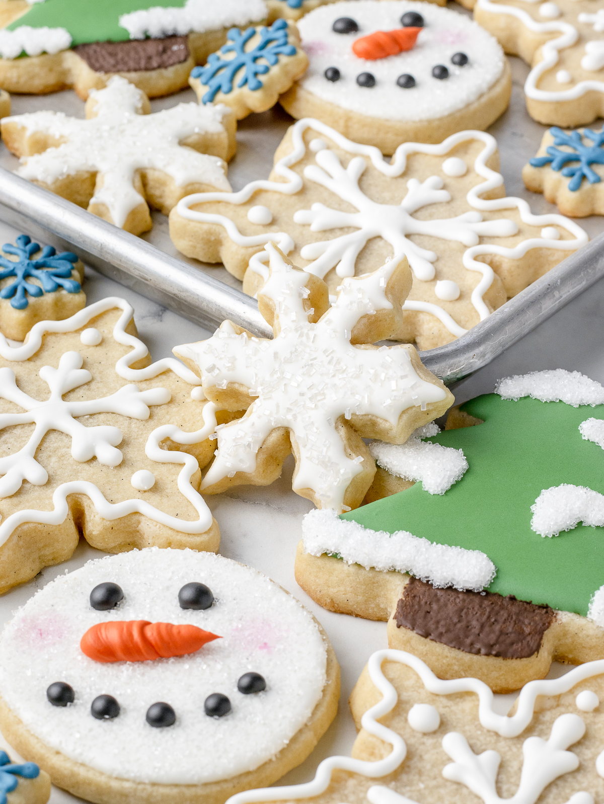 Different types of decorated Vanilla Sugar Cookies. There are snowflakes iced with sprinkles, pine trees with snow, and cute snowman faces.