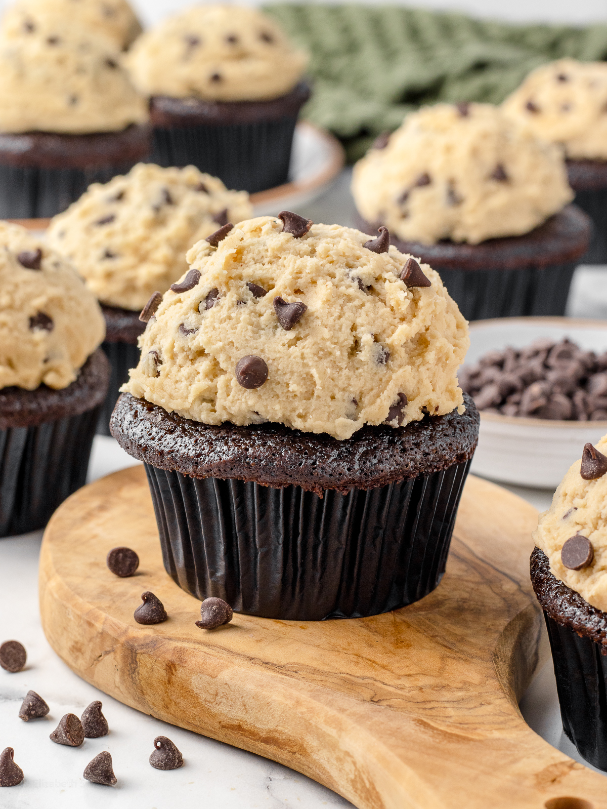 A bunch of Chocolate Chip Cookie Dough Cupcakes scattered around with mini semi-sweet chocolate chips on the side.