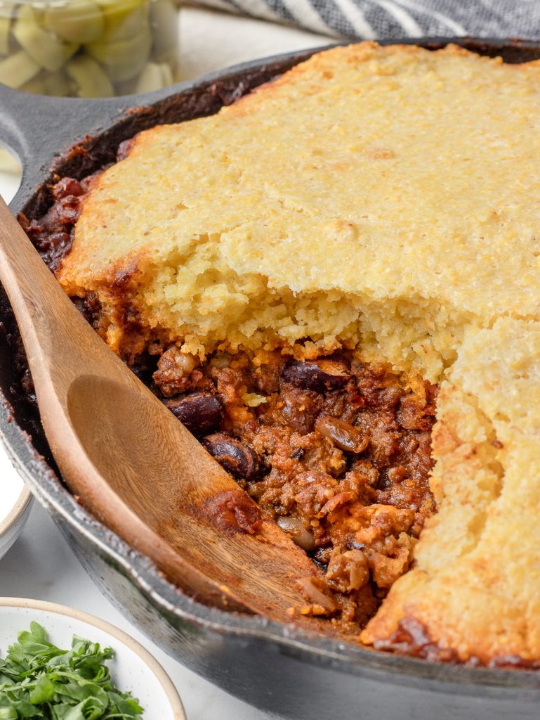 Spoon in a cast iron skillet of chili topped with cornbread. There is sour cream, cilantro, and pickled jalapenos on the side for serving.