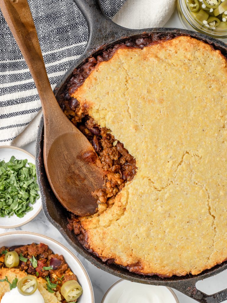 Spoon sitting in the Cornbread Chili Skillet. There is a bowl of it on the side along with sour cream, cilantro, and pickled jalapenos on the side for serving.