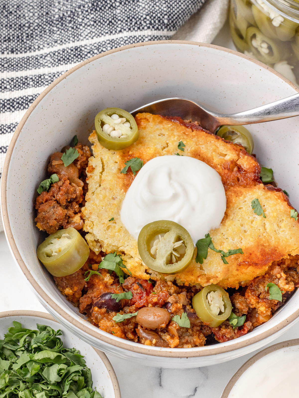 Big bowl of chili topped with cornbread, sour cream, pickled jalapenos and cilantro.