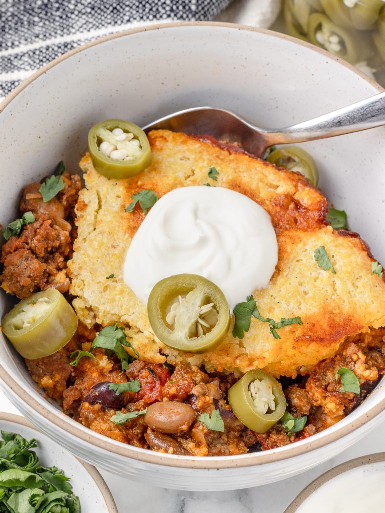Big bowl of the Chili Cornbread Skillet topped with sour cream, pickled jalapenos and cilantro. There is a spoon in it ready for eating.